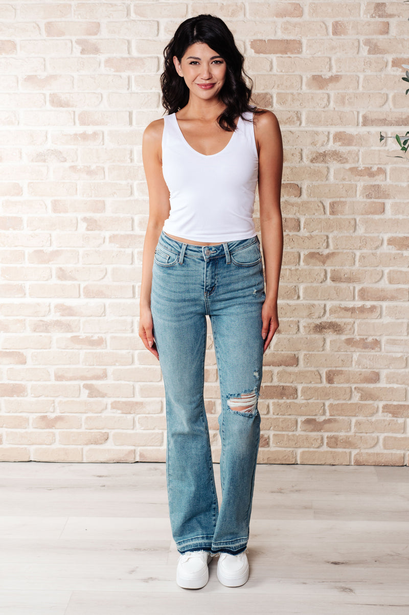 Judy Blue Mid Rise Bootcut Jeans. Designed with a mid rise, distressing at the knee, bootcut silhouette, and release hem detail with raw edge.